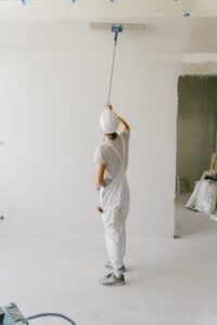 Back view of a painter in overalls and hard hat painting an interior wall with a roller.